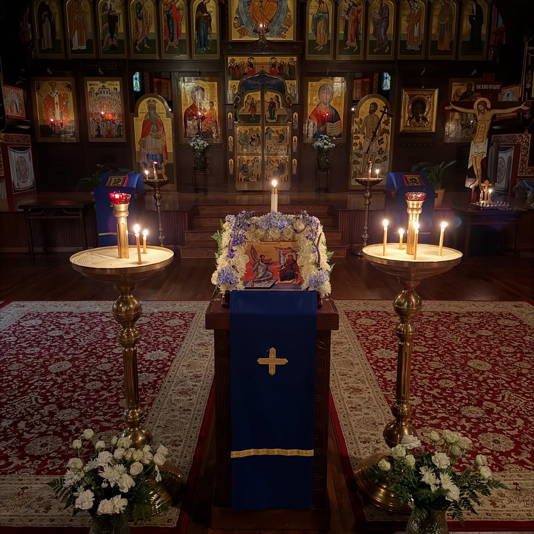 An icon on a stand draped in blue and framed with white and purple flowers, between two tall brass candle stands with lit candles, before the iconostasis
